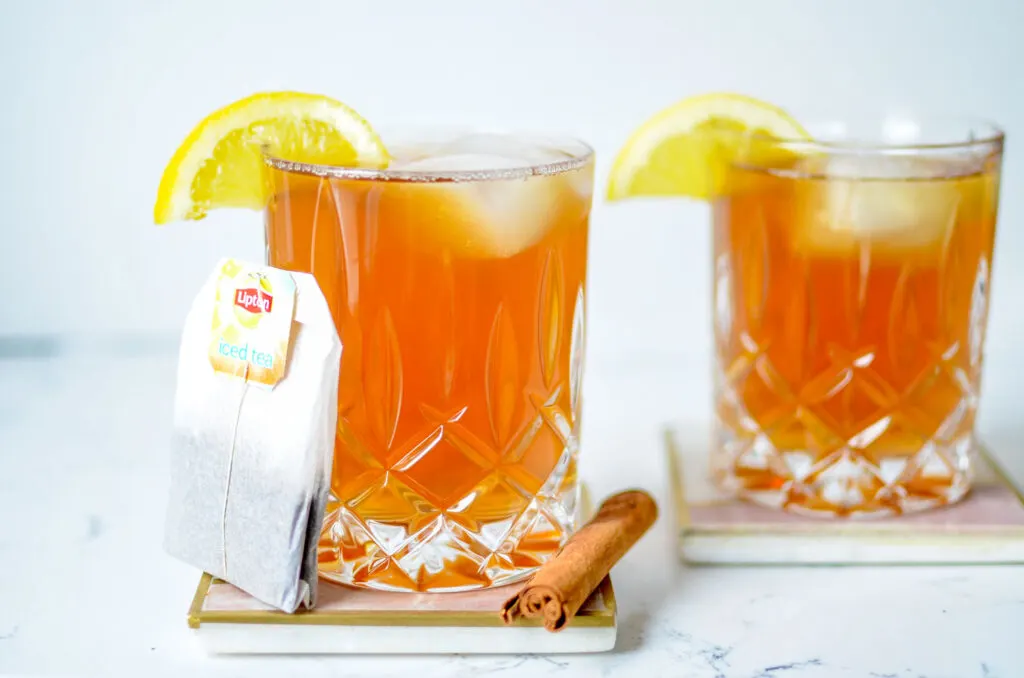 not-so-hot-hot-toddy-iced-tea-drinks-with-alcohol: 2 whiskey glasses on coasters on the counter. They're filled with an orange drink with ice and a lemon slice on the rim. There's a large tea bag leaning on the glass in front and a cinnamon stick on the side.
