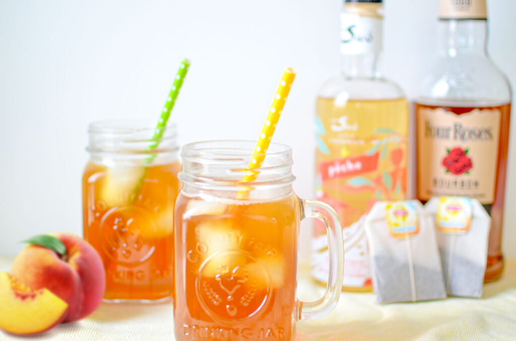two peach-iced-tea-cocktails in mason jars with handles and colourful polka dot straws and a peach next to them. Blurred in the background you see the peach liqueur, bourbon, and two large tea bags