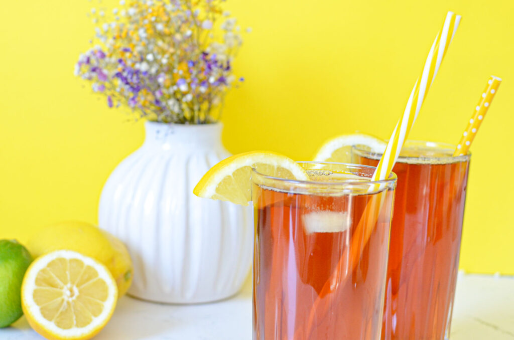 two-cups-of-ice-tea-with-a-yellow straw sticking out and slices of lemon the rims. its on a bright yellow background with a flower teapot with purple, yellow and white flowers in the background.