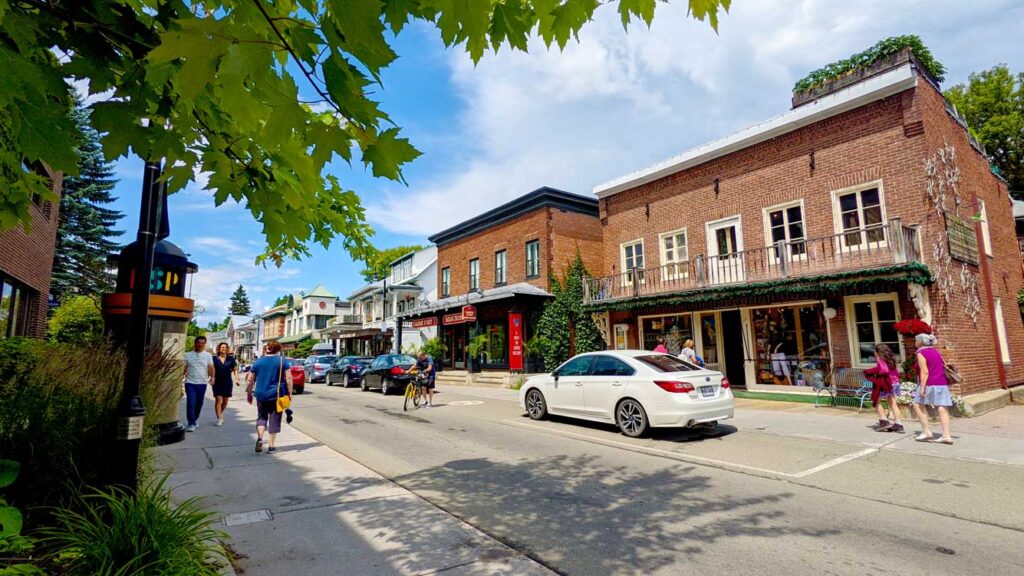 baie-sainte-paul-quebec bustling street in the summer with people walking on the sidewalks, small brick buildings housing shops, and cars driving down the middle on a beautiful sunny summer day