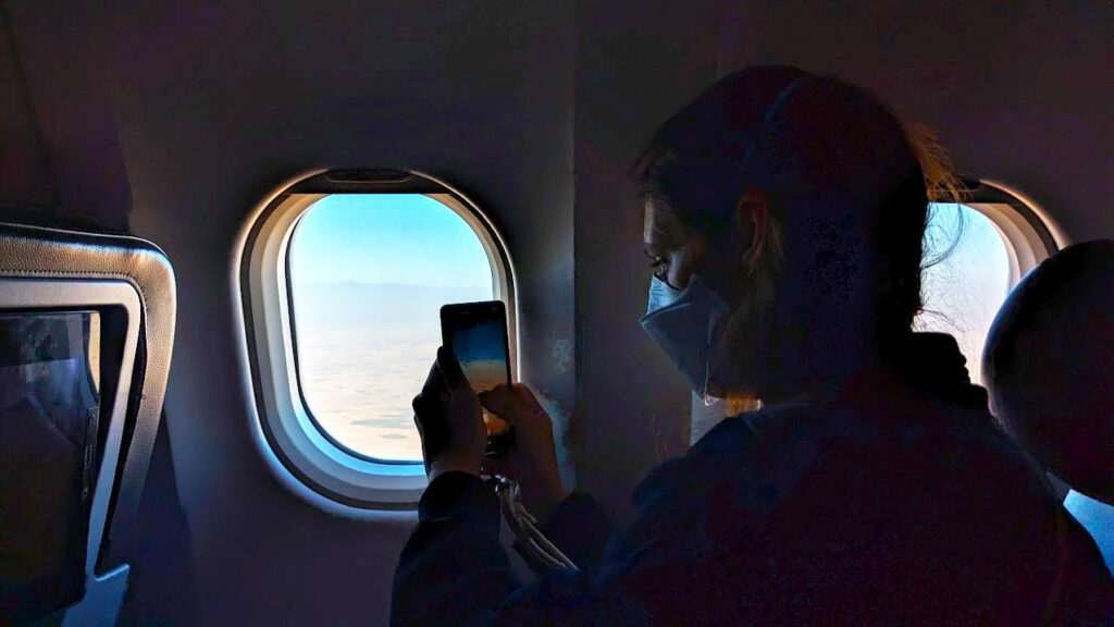 a pretty dark image. The only light is coming from a plane window, outside you see a blue sky and a reddish brown ground. The silhouette of a woman sits, looking out the window, she's holding her phone to photograph outside. You can't see her face because she has a white mask covering her mouth and nose. 