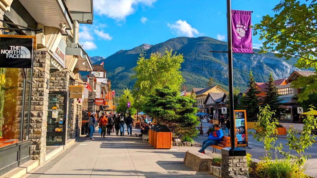 the streets of banff avenue in banff town national park canada on a very sunny day. A shop is on the left side, with trees lining the other side of the sidewalk. And a glorious canadian rocky mountain is in the background