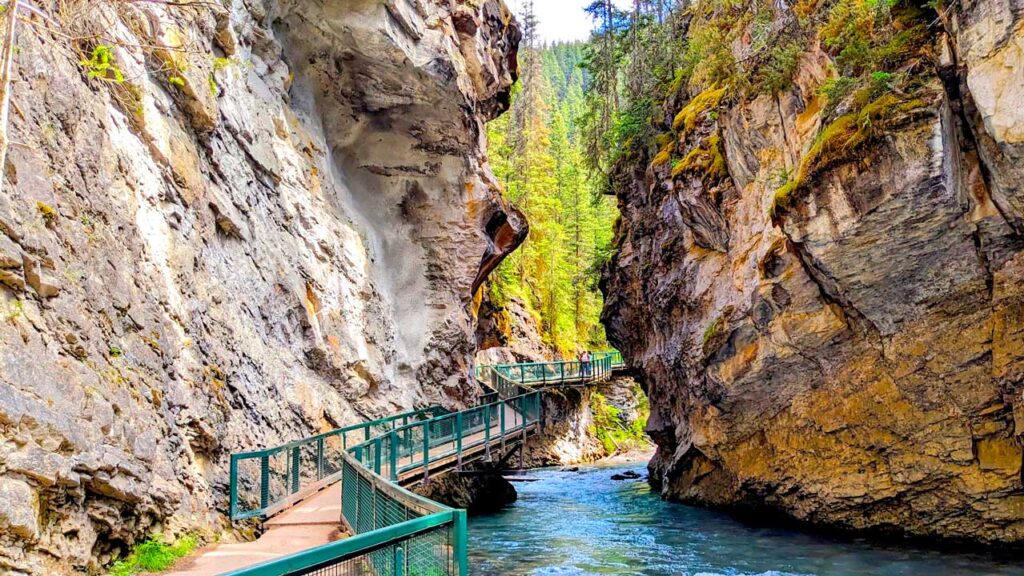 johnston canyon suspension bridge is attached to a rocky cliffside hanging above the rushing teal glacier water below. It wraps all the way around until you can't see it anymore. On the other side of the water is another rock wall. You can't see the top of the walls, but through them above the water is a layer of green trees. 