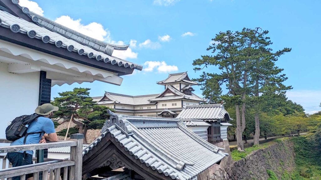 a man standing inside kanazawa castle grounds, on a raised outlooking, looking at kanazawa castle in the distanced. It's an iconic Japanese architecture roof and build, it's white and grey. There's a tree next to the main building. And you can see the stone wall separating the castle grounds from the park surrounding it