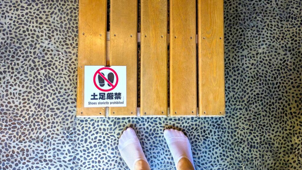 a top down view of a pebble filled floor, and a raised wooden floor on top. On the wood floor is a sign that says in both Japanese and English: shoes strictly prohibited with a visual sign as well. In front of the wood floor is a pair of feet wearing thin white ankle socks