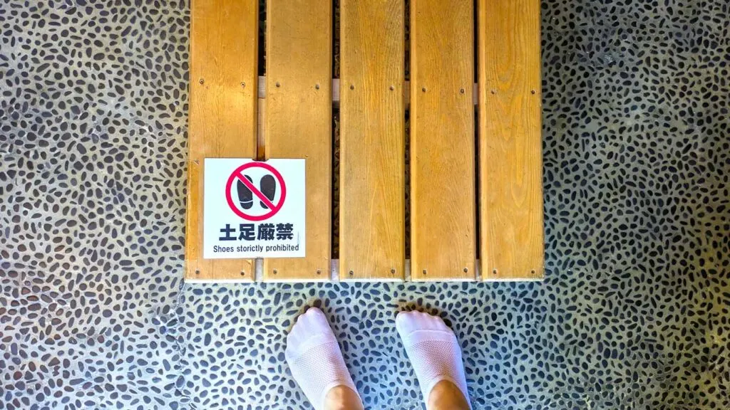 a top down view of a pebble filled floor, and a raised wooden floor on top. On the wood floor is a sign that says in both Japanese and English: shoes strictly prohibited with a visual sign as well. In front of the wood floor is a pair of feet wearing thin white ankle socks