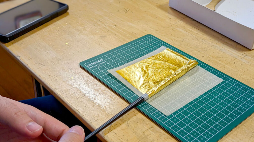 gold leaf experience in in kanazawa japan, making your own gold leaf chop stick. A very very thin sheet of gold rests on an art cutting board, it's being rolled into a chopstick, covering the top half. 