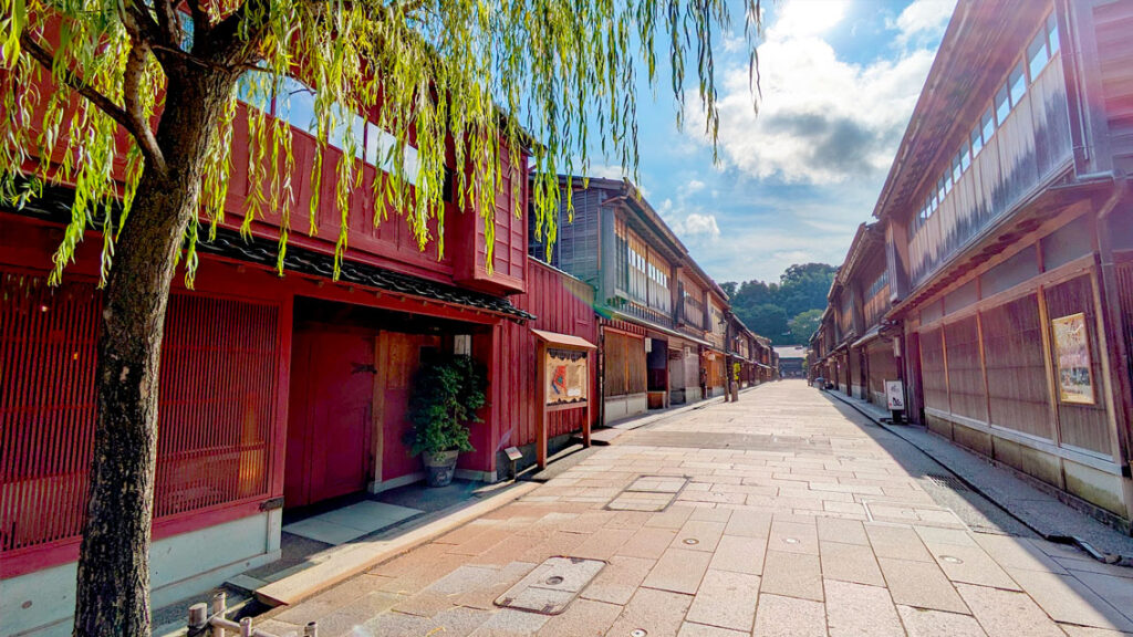 higashiyama ward old tea district in kanazawa japan. Stone street with old red wood buildings lining the street. A tree with droopy leaves frames the top left side of the image, adding a vibrant pop of green to the photo. Not a single person is walking in the streets, it's empty!