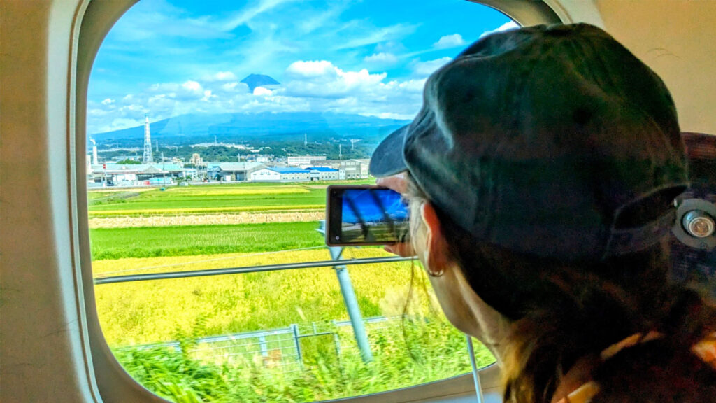 seeing mount fuji from the shinkansen. You can barely see that you are looking through a window seeing the edges of a frame. But the back of a womans head blocks the right side, she's holding a phone in her hand, photographing what's outside: across a field, with some clouds covering the middle, is the iconic mount fuji