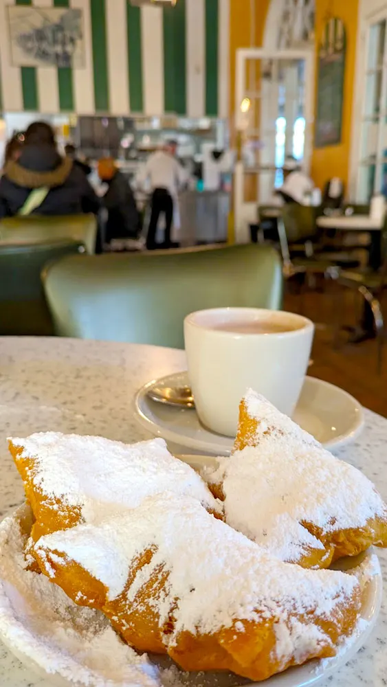 three giant beignets (squared donuts with no holes, and COVERED in powdered sugar, they're more white than fried dough) sit on a small white plate on a round table. Behind them is a matching simple white mug, filled with cafe au lait. You can see the rest of the restaurant, an old style design, because it hasn't been updated in years. The floors are a dark wood , and it has green and white vertical striped wallpaper