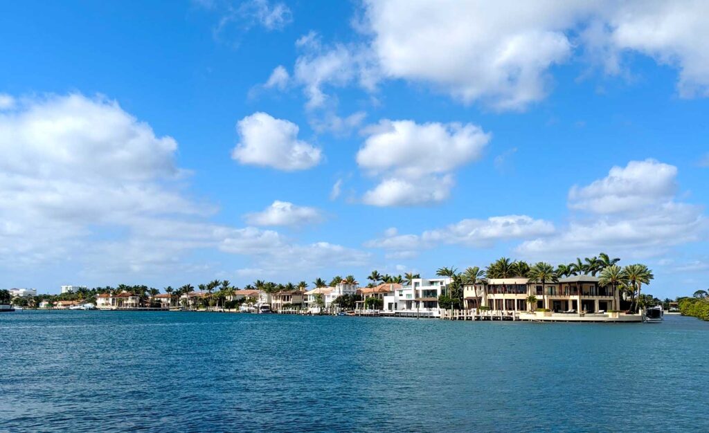 the view from across the water, there's a small sliver of land separating the rippled ocean from the cloud spotted sky. The land is filled with mansions, with barely room between them for a palm tree. They look like villas with patios, and pastel coloured.