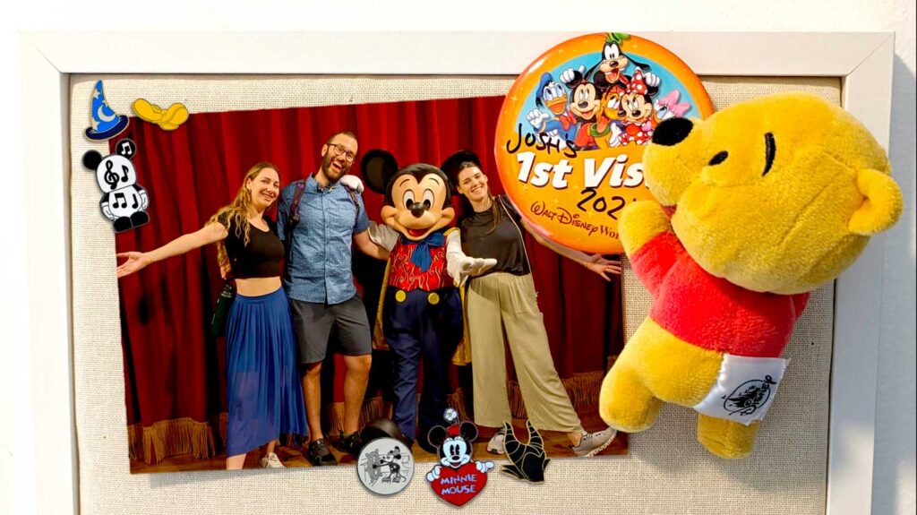 what-you-can-do-with-disney-collectible-pins-at-home-a-closeup-of-a-corkboard-with-a-picture-of-three-adults-with-mickey-mouse-holding-up-the-picture-are-several-disney-pins-a-first-time-occasion-pin-and-winnie-the-pooh-shoulder-plush