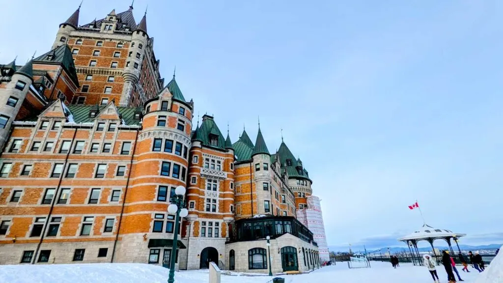 chateau frontenac towering above place duffering covered in snow below. There's a small tent with a flag on it for a size comparison. The hotel is beautiful stone and red brick design with spike green roof towers 