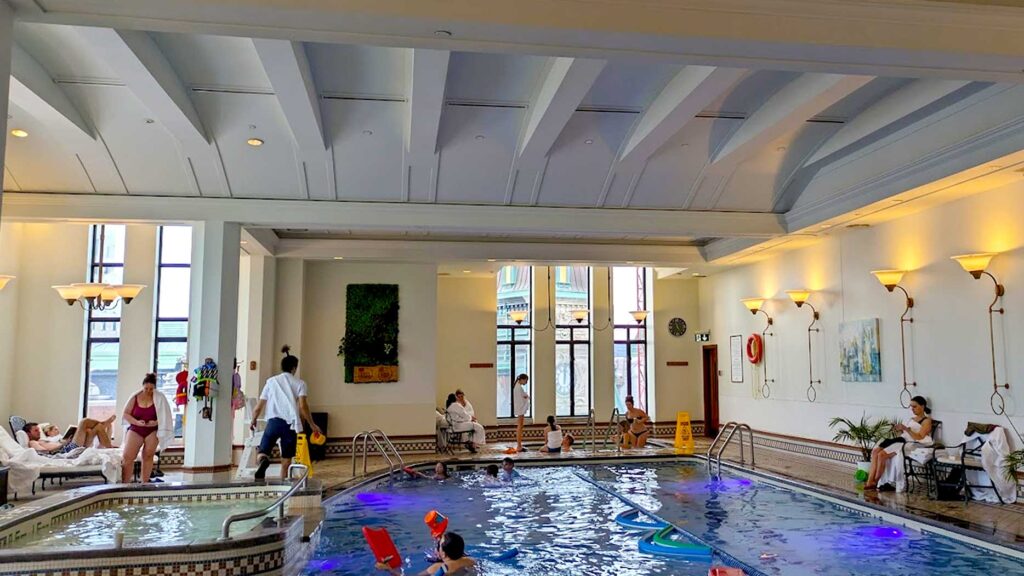 chateau-frontenac-pool-quebec-city that a lot of families are enjoying. Theres a regular large pool with kids and parents in it and an empty hot tub next to it. Adults are reading books on the lounge chairs, while others are watching their kids in the pool