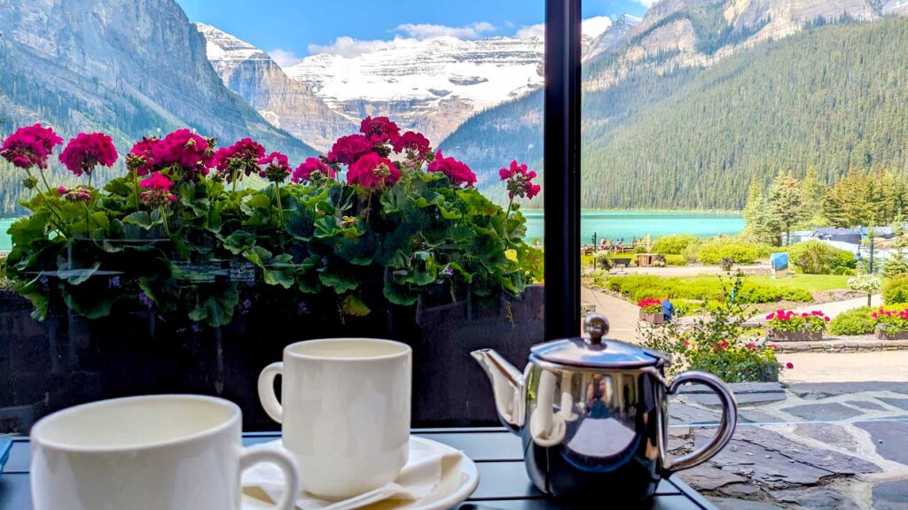 chateau lake Louise tea at fairview restaurant, sitting at an outdoor table, two white tea cups and a silver teapot . Theres a planter behind it with bright pink flowers. Behind that you see the priceless lake Louise view. Giant snow capped mountain on bright blue glacier water