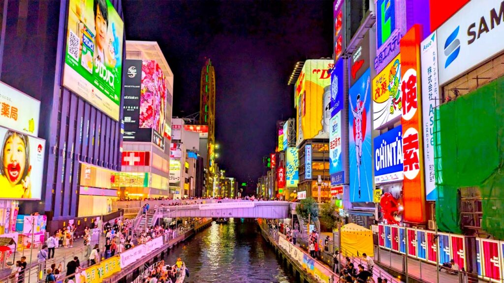 dotonbori district at night. standing on a bridge in the middle of the canal. the sky is pitch black but the canal sidewalks and bridge are lit up by the larger than life neon signs on the buildings lining the canal. The sidewalks are PACKED with people enjoying the views.