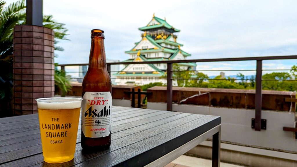 enjoying an asahi beer outside on a rooftop terrace. the beer bottle with a plastic cup filled with beer next to it. You can see a blurred osaka castle in the background with it's white temple and green roofs with golden decor