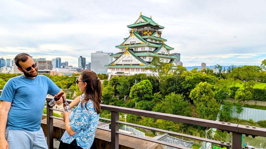 two people standing on a rooftop terrace each with an asahi beer bottle in their hand, leaning on a railing. In the background behind them is the towering osaka castle with white, green and gold accents. It towers above the trees around it and buildings far off in the background behind it