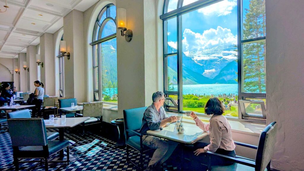 a couple seated by the exclusive seats by the window with a teacup in their hand lookout out through the giant rounded window at the view of lake louise. You can see the empty seats all around them in the rest of the grand room