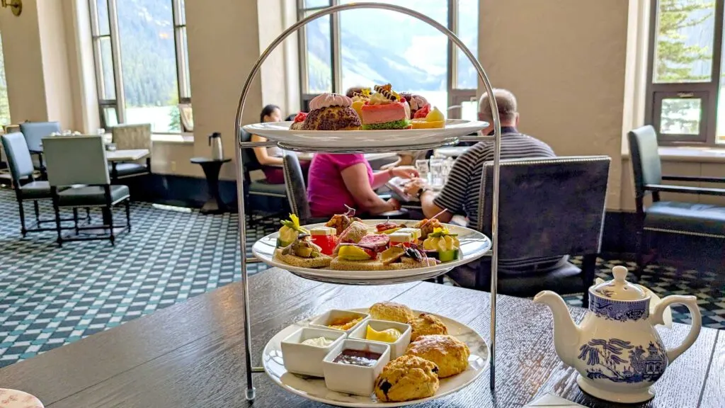 fairmont high tea at the chateau lake louise. seated at a table in the third row, you can see two rows of tables in front with large rounded windows behind. A lot of light is coming in, the outside is almost too white to see but you can slightly make out the mountains and Lake Louise. On the table is a three tiered afternoon tea plate filled with small desserts, tiny sandwiches and 4 large scones on the bottom. There's a blue and white victorian style teapot.