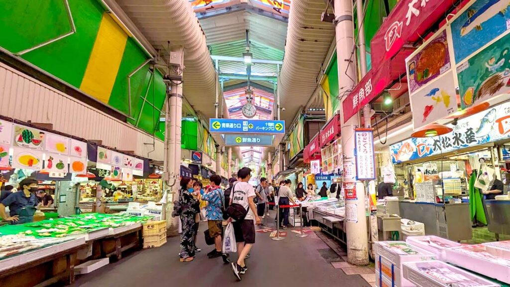 a food market in japan. on the left is a fish vendor with dead fish laying on the shelf of the food star being displayed to people walking by