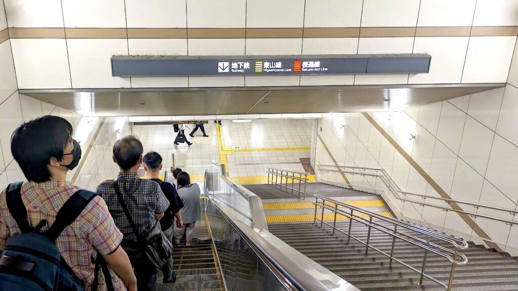 people standing in a single file line on the left side of an escalator going down to a subway in japan. There are signs on the ceiling in front directing you which way to go, in japanese. And there are completely empty stairs next to the escalator