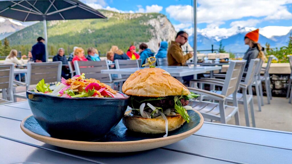 an outdoor terrace surrounded by mountains and blue skies. On the table is a burger with a salad