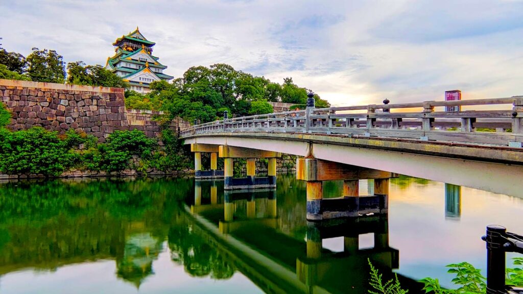 osaka castle across the moat leaving a reflection on the water below. its golden hour leaving a pinkish, blueish, yellow hue. a wooden bridge is leading you across the water below.