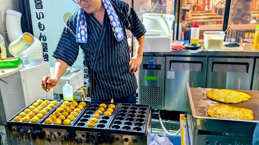 a street vendor in japan standing in their food stall making takoyaki balls on one side, in the process of flipping the balls over to cook evenly. On their left is another flat stove top where two okonomiyakis are cooking