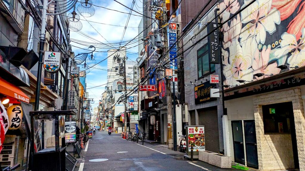 a view of the deserted streets in osaka. no one is there. but yet the street is overwhelmingly full. theres barely a sidewalk to walk on. The walls of the buildings are filled with ads and signs. You can barely even see the sky through all the wiring crossing the street. 