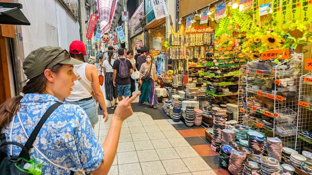 a woman with a surprised look is point her finger at one of the many shops down the street. There are a bunch plates on the floor for sale among other kitchen supplies in the front of the store

