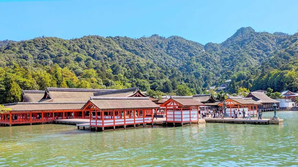 Itsukushima Jinja Shrine. a floating shrine. Several interconnected docks with rooftops for shade. The pillars are painted red to match the otorii gate. It looks like a village floating on water. Behind the village is a mountain filled with trees