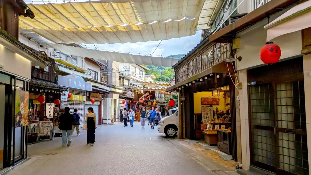 Omotesando Shopping street miyajima island. Both sides of the street are lined with vendors, all the buildings are attached to each other all the way down the street, curving with the street. People are walking in the distance, going in and out of the shops. 
The stores on either side are attached to each other by a fabric tarp over the top, to protect from the summer heat. There are breaks between tarps, where you can see some clouds in the sky .