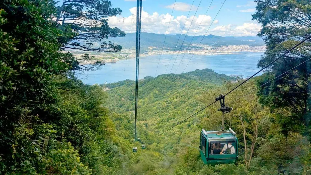 the view from the first cable car a part of the miyajima ropeway. You're looking down on the mountain below, across from you is another green cable car making their way up the mountain instead. A rope is directly above, leaving the frame in the middle, but it goes all the way down disappearing somewhere in the middle of the photo, in all the trees below. Beyond the tree filled mountain, you can see the sea, and the land across from it. There is a city on the other side of the water, but there's nothing distinguishable. Behind the city is more mountains