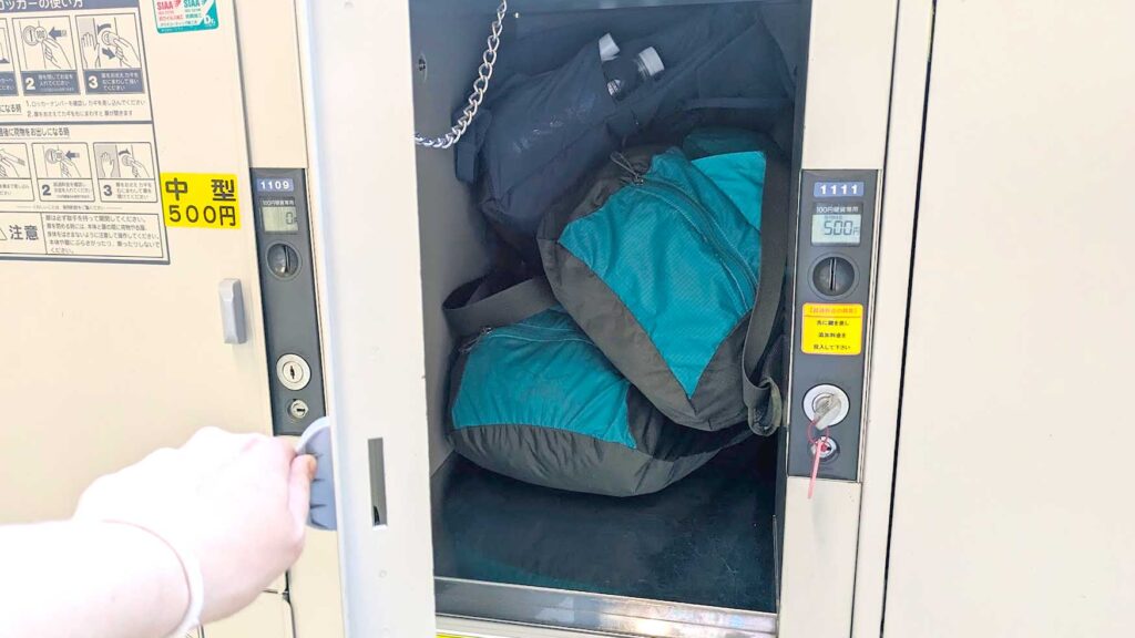 a hand holding the door open to a coin locker in japan. Inside the coin locker are two packable duffel bags and a backpack