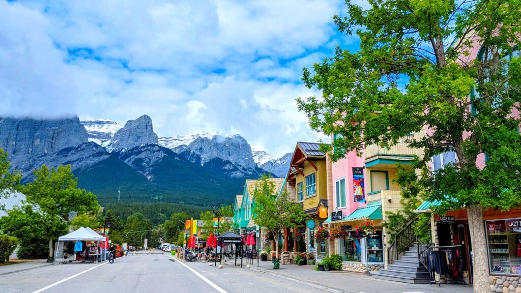 colourful homes line the pedestrian only 8th street in canmore alberta. There's not a car in site, but people sitting on the terraces for the restaurants. The buildings are painted such colourful colours like yellow, pink, green and red. Behind the street are the towering mountains of the Canadian rockies.