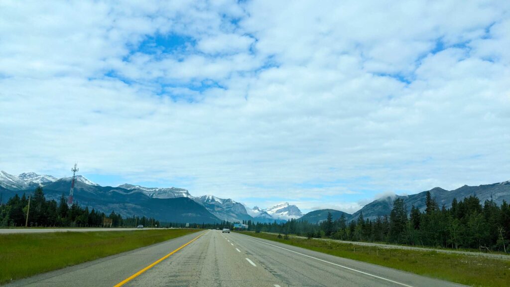 the long road forward leading you towards the canadian rockies in the distance. Even though it looks like summer where you are with green trees and grass, the mountains in the distance are still snow capped. 