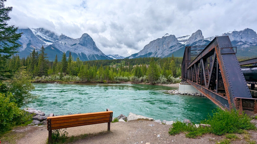 rushing teal coloured water of Bow river is surrounded by evergreen trees on either side. Closest to you is a bridge to sit on and enjoy the view across the river of the rocky mountains. On the right of the frame you see a large iron bridge, engine bridge, to cross the river safely. 