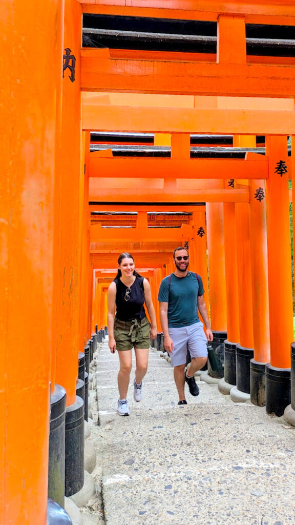 a couple are walking through the fushimi inari shrine on an easy day trip from kyoto japan. the up hill path is finished, long stone steps. Lining the path are bright orange tori gates, there's barely a gap between each one. The couple walks forward as they make their way up the hill