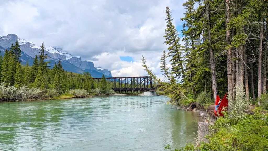 the most relaxing view of the bow river in canmore alberta. Mount Rundle lines the frame leading your eye from the left to the distance, where you meet engine bridge, a brown rustic, and very practical looking bridge taking you across the rushing teal water below and onto the other side of the river. On this side, tall trees line your view. In between the tall trees, you can see something red, it's someone sleeping in a bright red hammock.