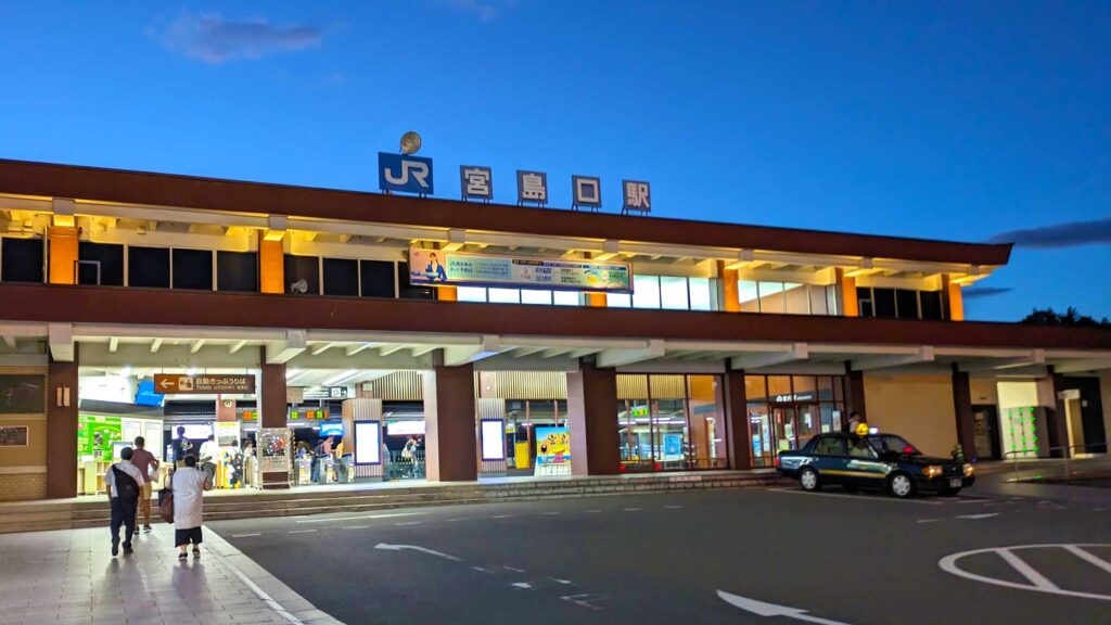 the outside of a jr station at dusk. There's a JR sign on top of the one story station. People are walking in to the entrance. There's a taxi parked waiting outside for anyone who needs it. 
The lights are on inside the station. Adding a wonderful colour palette to the photo. The pale blue to royal blue from the sky pairs with the orange to yellow lights . 