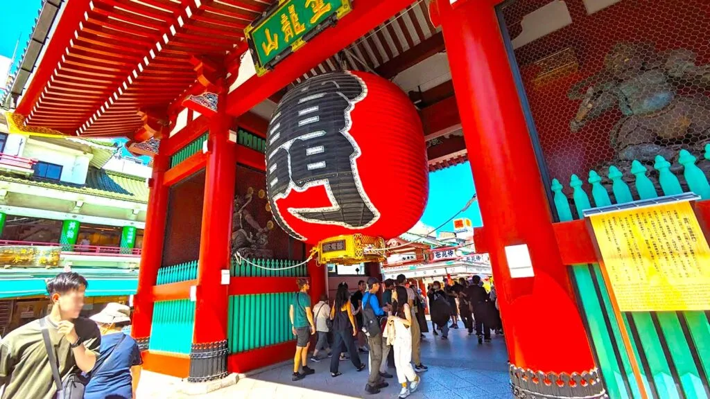 kaminarimon giant lantern at the senso-ji shrine in tokyo japan. People are crowded underneath, you can barely make it through. The lantern is red with kanji printed on it in black. It hands from the shrine gates, that are also painted a bright red
