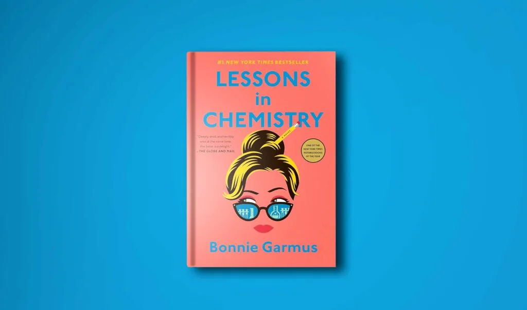 hardcover book on a plane blue background to compliment the orange book cover of lessons in chemistry by Bonnie Garmus