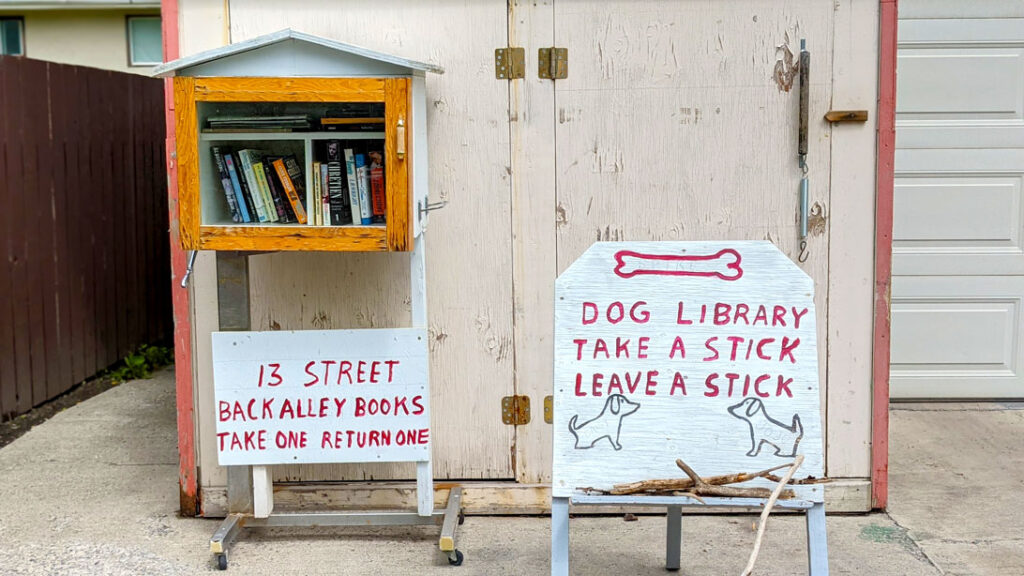 an example of local vibes in canmore alberta. Outside a house are two signs. One is attached to a mini raised house, with a glass door, showing off some books inside. The attached sign says:  13 Street Back Alley Books, Take one Return one. 

Next to it is a larger sign with the painted outline of a dog bone, it reads below: dog library. Take a stick, Leave a stick. There are two outlines of sitting dogs looking at each other below the text.