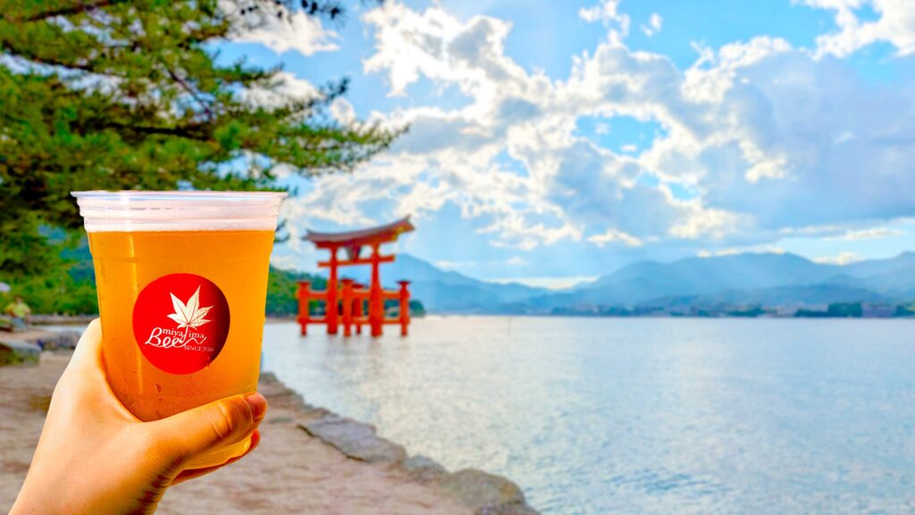 a hand coming in to the left of the frame holding up a light beer from Miyajima brewery. The sunlight is hitting it lighting it up. In the background is slightly blurred but you can still clearly see the floating torii gate, a bright red, rising from the water below. Behind it are the sloping mountains and the sun is peaking through the clouds. 