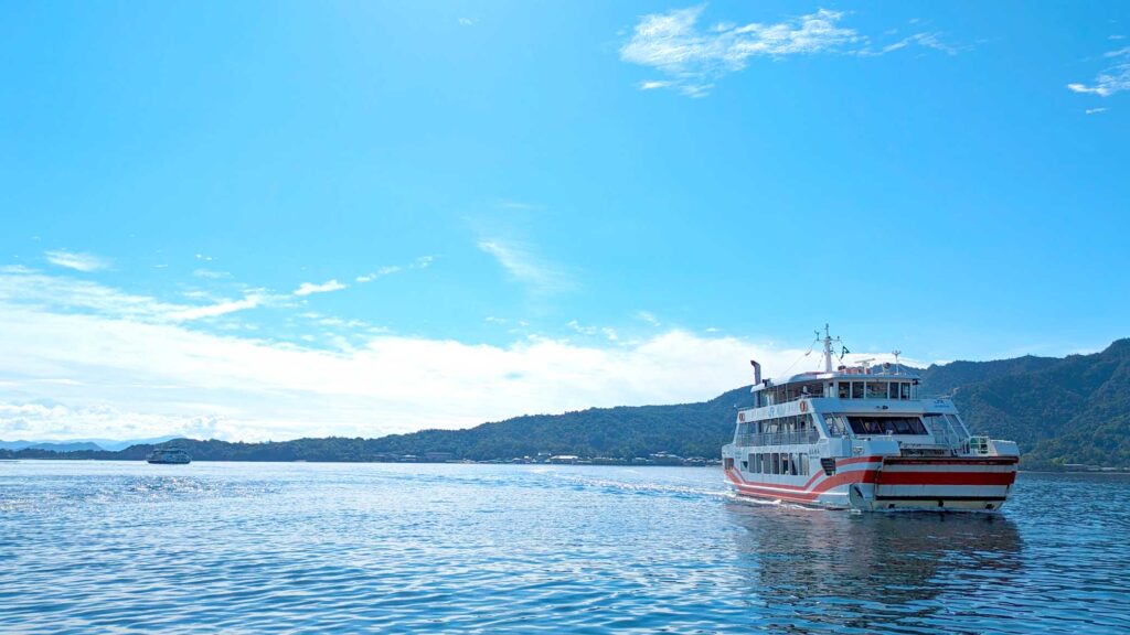 a very simple photo of land sea and sky with a red and white miyajima JR island ferry coming in towards the land on the right of the photo. 