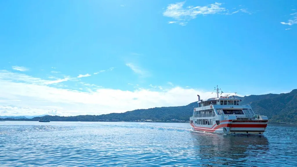 a very simple photo of land sea and sky with a red and white miyajima JR island ferry coming in towards the land on the right of the photo. 