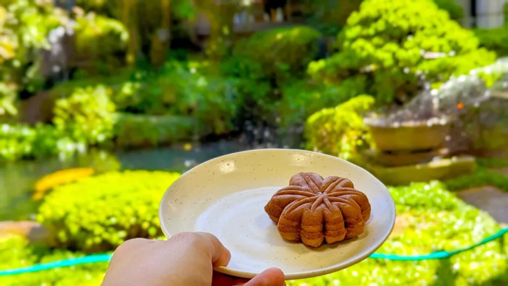 a person holding up a plate with a maple leaf dessert . behind it is a bright sunny Japanese garden with a pond and lily pads and lush green bushes. 