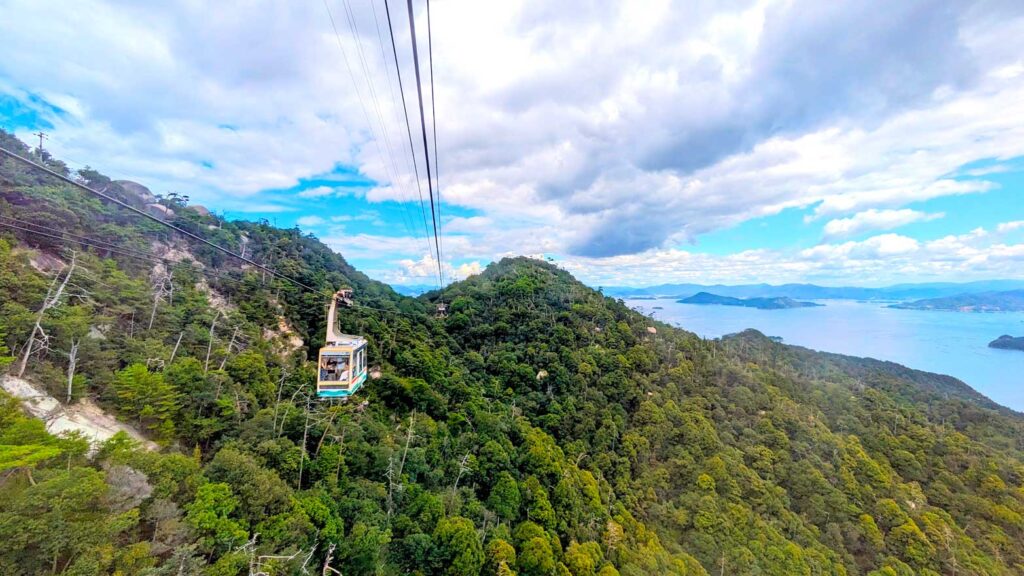 the view from the miyajima ropeway. you see the large cable car fitting about 15 people hanging on a rope high above the trees below making it's way towards you. Beyond the trees, you know you're high up because the sea is off in the distance behind the mountains