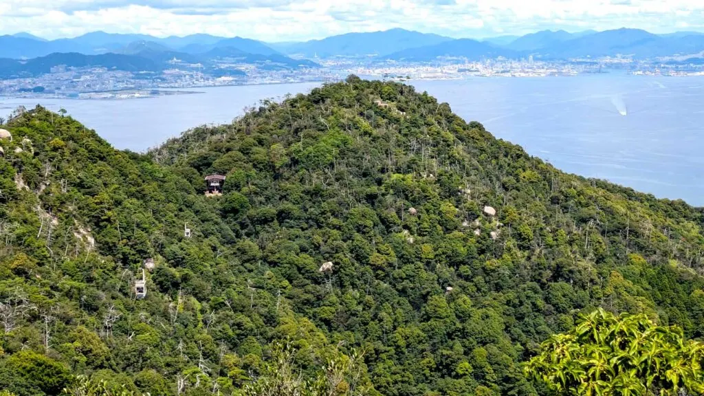 two peaks of a mountain range below you, you can slightly make out two rapes and cables cars dangling from them. Behind the mountain is the sea and a city on the land with even more mountains behind it
